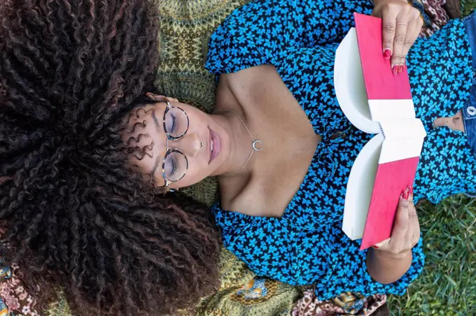 Afro young woman with curly hair reading book while lying down at public park