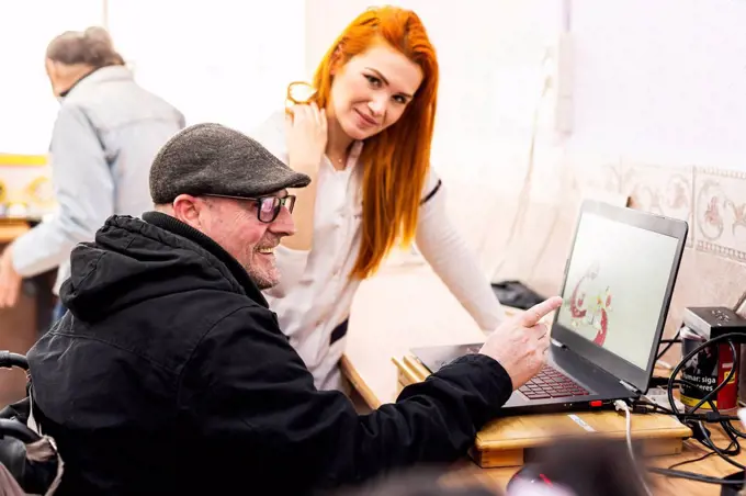 Smiling man using laptop by female caregiver in disabled center