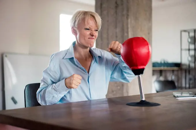 Blond businesswoman punching boxing bag on table at home office