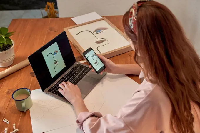 Female artist sharing photograph of drawing online through laptop at desk in home