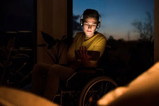 Young man in wheelchair wearing wireless headphones while using digital tablet sitting on wheelchair in living room