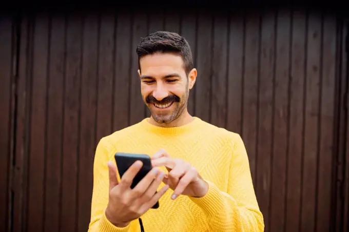 Smiling mid adult man using mobile phone against wooden wall
