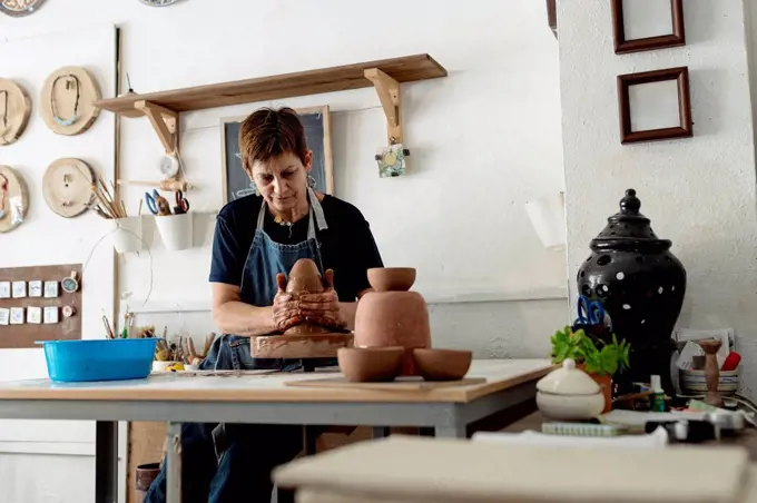 Mature female artist making pottery from brown clay in ceramic store