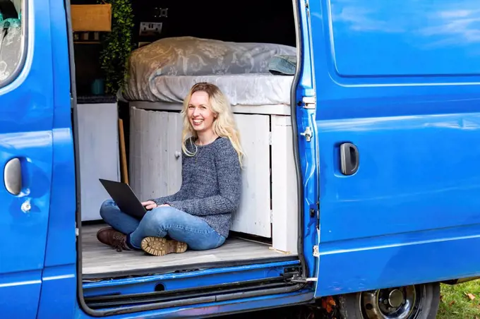 Happy woman with laptop sitting at door of camper van