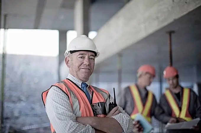 Confident male contractor with arms crossed at site with construction workers seen in background