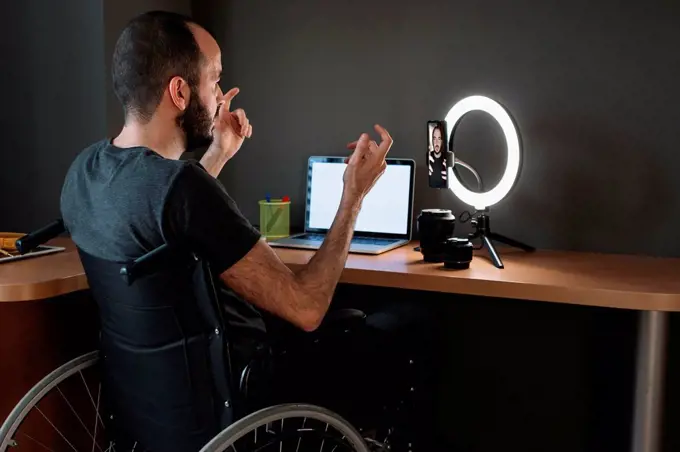 Vlogger filming through mobile phone with circular strobe tripod while sitting on wheelchair at studio