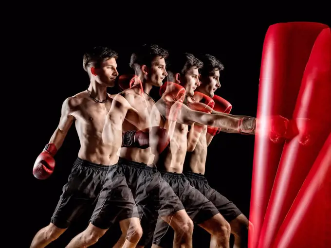 Young male boxer punching red bag against black background