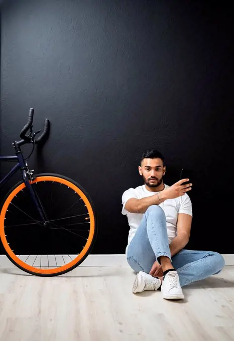 Young man with smart phone sitting near bicycle by wall at studio