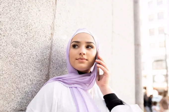 Arab teenage girl looking away while talking on smart phone by wall