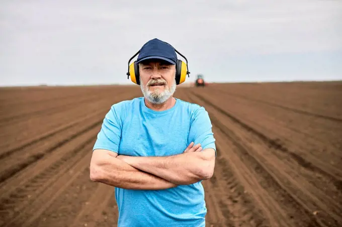 Male farmer wearing ear protectors while standing with arms crossed on agricultural field