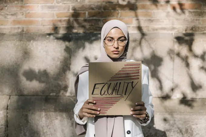 Young woman holding placard with equality text in front of wall on sunny day