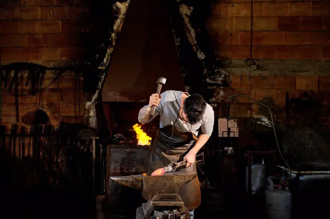 Male metal worker forging metal with hammer at blacksmith shop