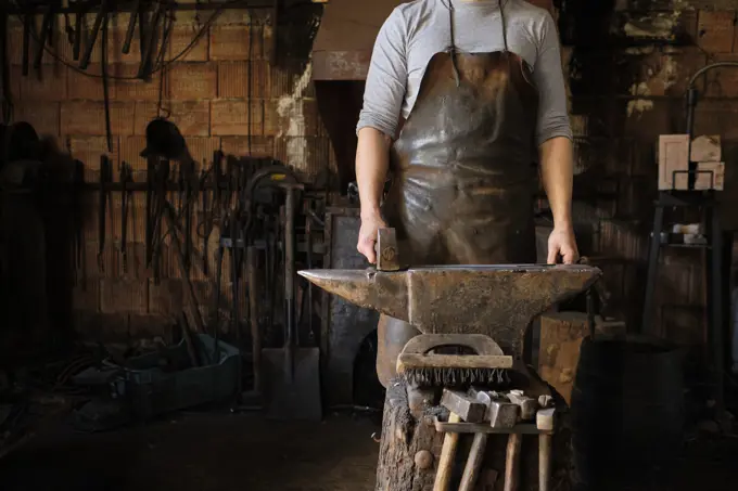 Male blacksmith standing with hammer by anvil at shop