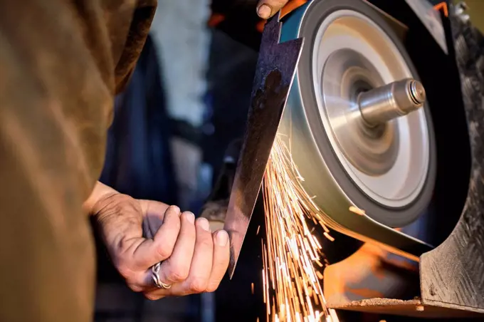 Male blacksmith sharpening knife shape metal on grinder at workshop
