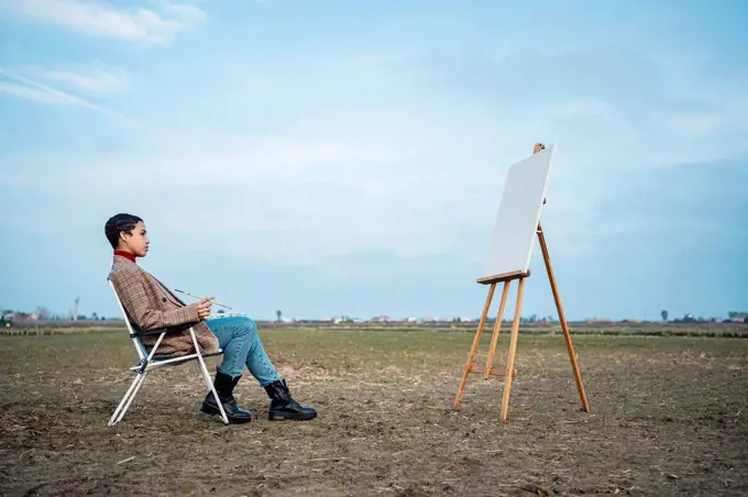 Thoughtful woman looking at easel while sitting on chair in countryside