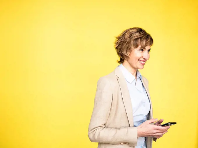Smiling businesswoman holding mobile phone by yellow background