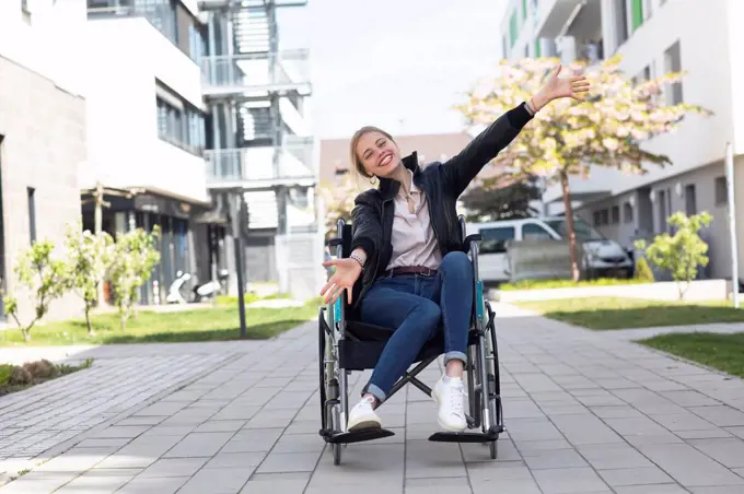 Happy disabled woman with arms outstretched sitting on wheelchair in residential area