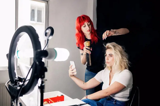 Female stylist applying spray on model's hair taking selfie through mobile phone in studio