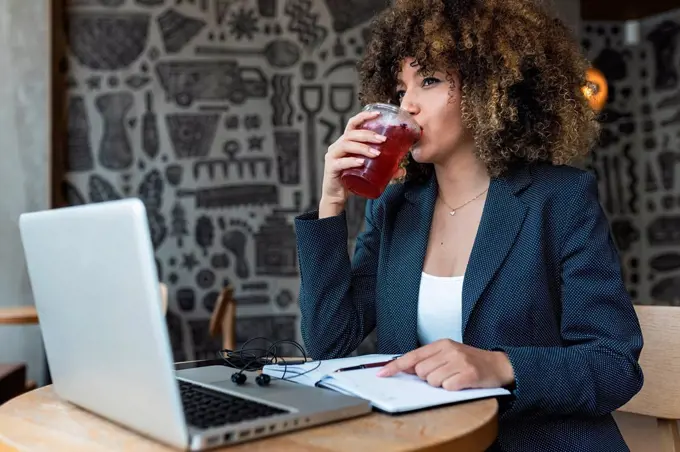 Afro businesswoman looking away while drinking in front of laptop