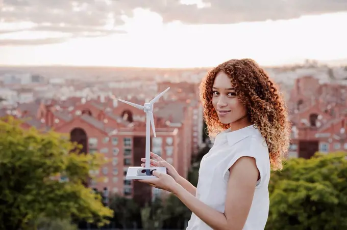 Young Hispanic woman holding wind turbine standing by city