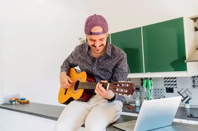 Smiling male composer playing guitar by laptop on kitchen island at home