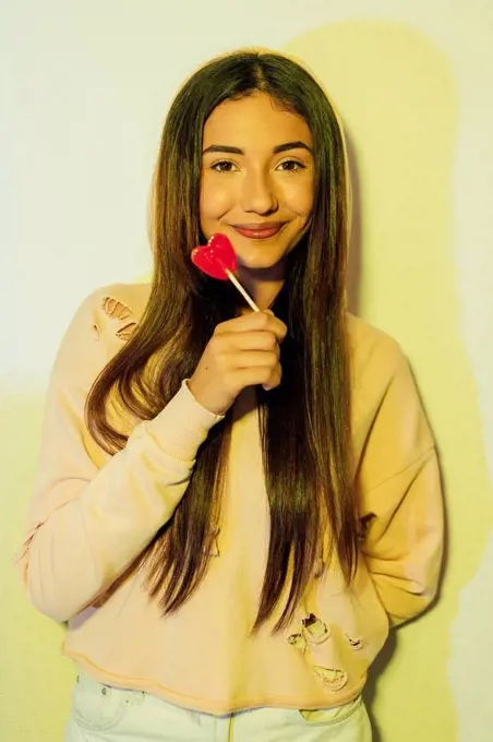 Beautiful woman holding heart shaped lollipop in front of wall