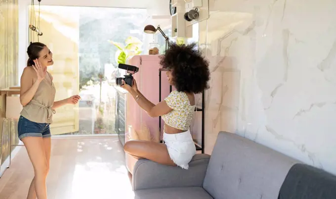 Young woman waving in camera held by afro female friend while filming at home