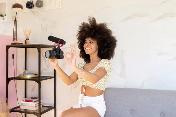 Afro woman gesturing while holding camera at home