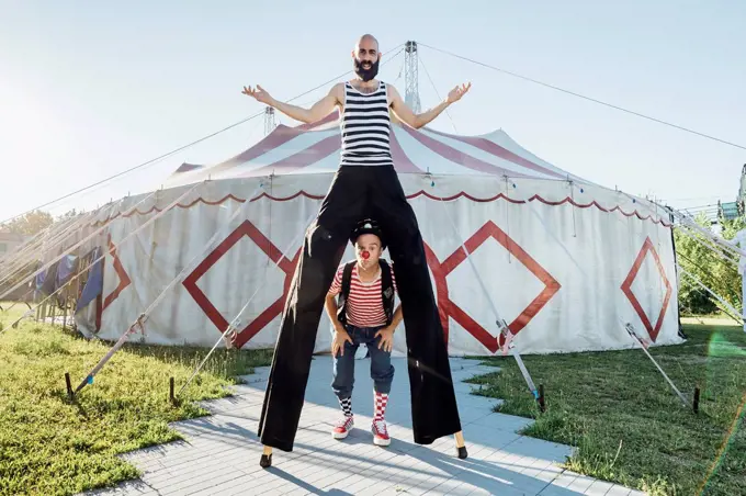 Male clown standing with performer on stilts in front of circus tent