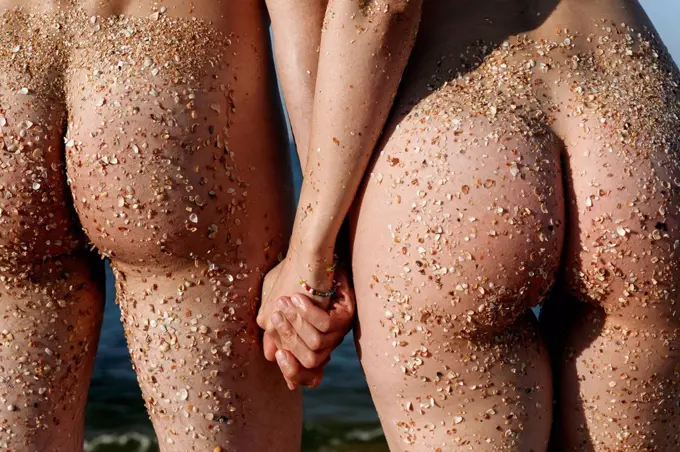 Naked couple with sand on buttocks holding hands while standing at beach