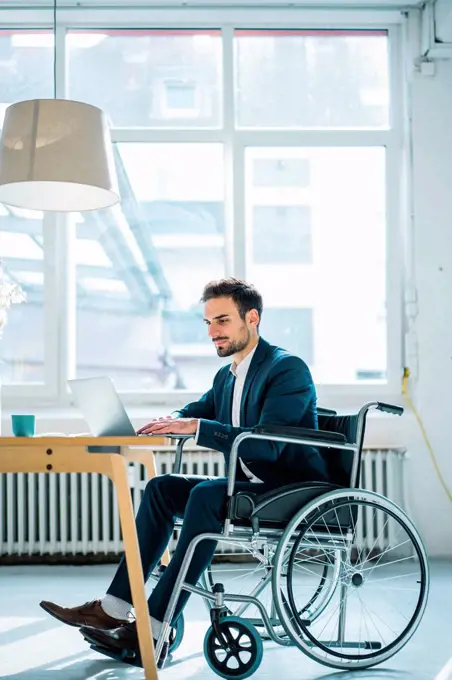 Young disabled male professional using laptop while sitting in wheelchair in office