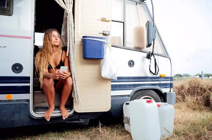Young woman holding smart phone while sitting at doorway of camping van