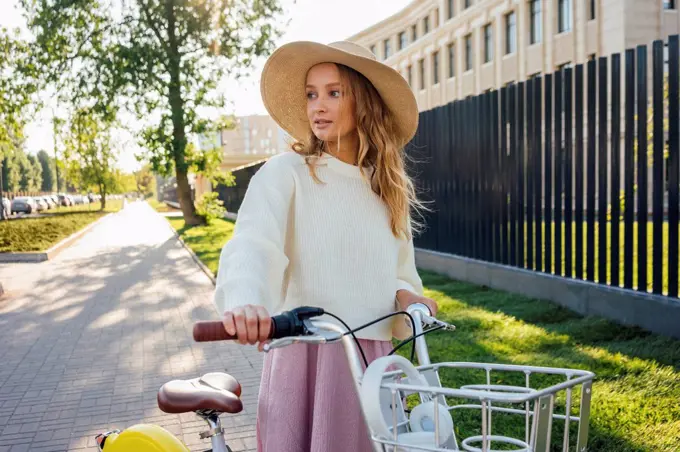 Young blond woman wearing hat standing with bicycle at park