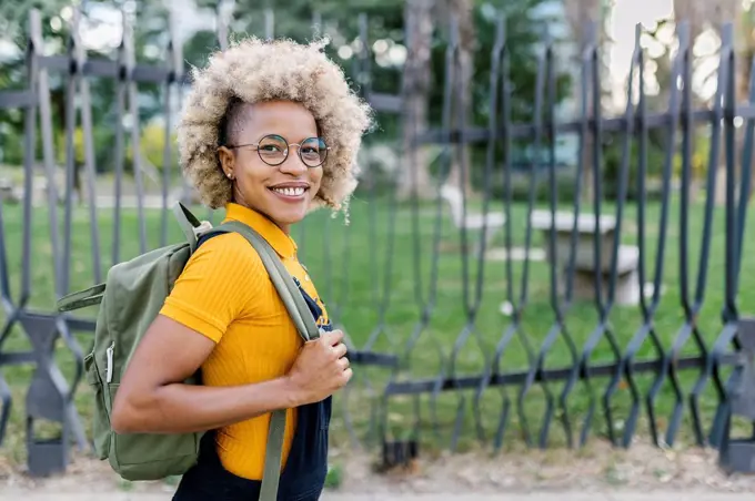 Smiling woman with eyeglasses carrying backpack by fence