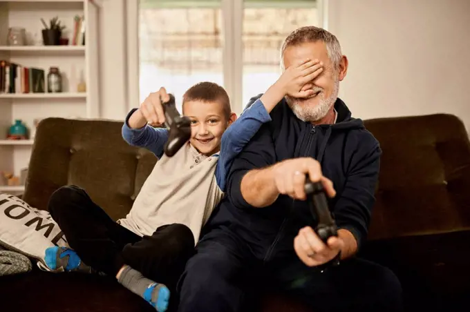 Grandson covering eyes of grandfather playing video game at home