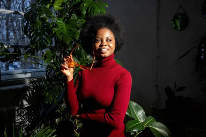 Afro young woman with sunglasses day dreaming by plants at home