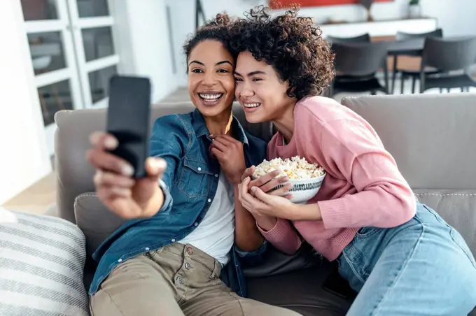 Happy friends taking selfie through mobile phone sitting on sofa in living room