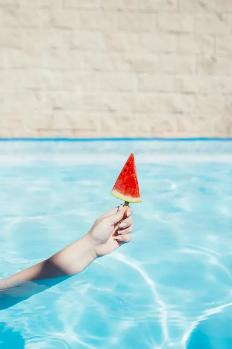 Hand of woman holding watermelon slice with popsicle stick in swimming pool