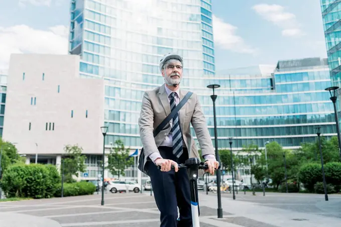Businessman riding electric push scooter in front of modern office buildings