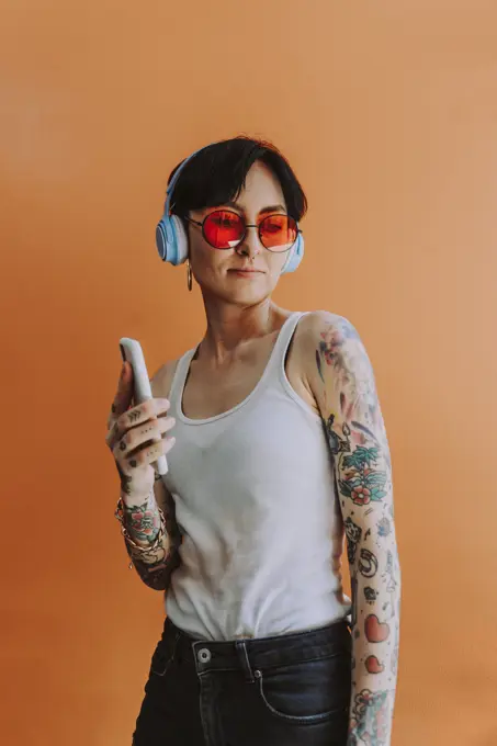 Beautiful woman listening music through wireless headphones standing against orange background