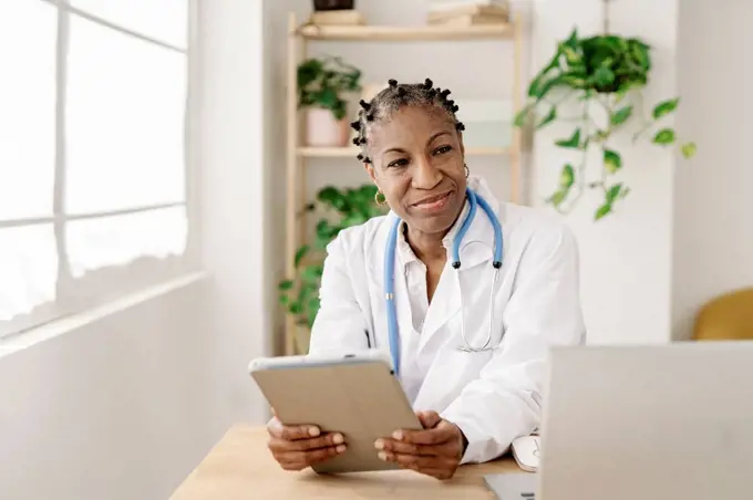 Smiling female doctor with tablet PC sitting at table in home office