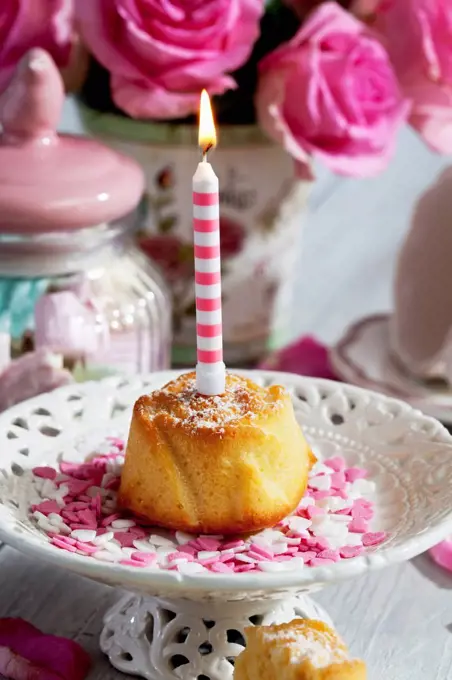 Muffin with lighted birthday candle, glass of marshmallows, cup and pink roses on table