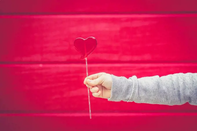 Close-up of boy holding heart-shaped lollipop in front of red wall