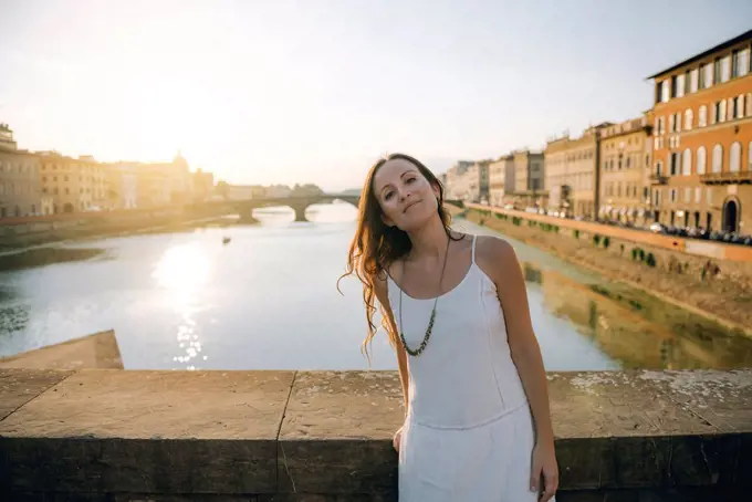 Italy, Florence, portrait of woman wearing white summer dress standing on a bridge at sunset