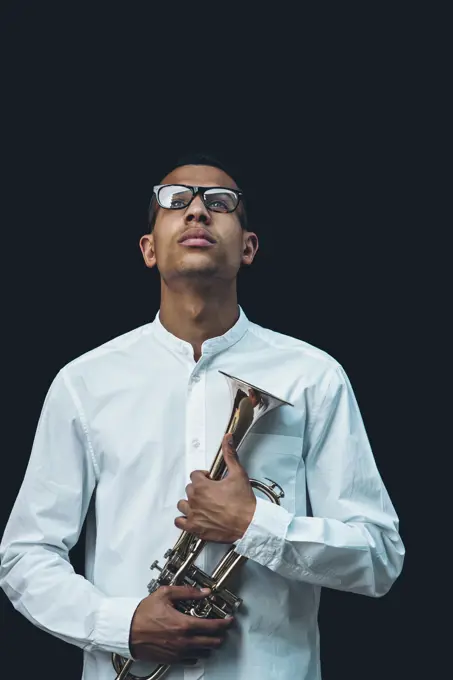 Portrait of young man wearing glasses and white shirt holding trumpet in front of black background