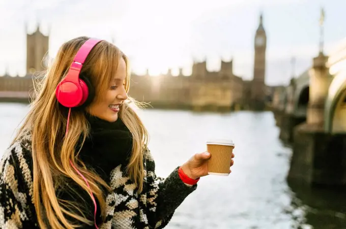 UK, London, young woman listening music and drinking coffee near Westminster Bridge