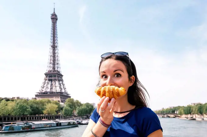France, Paris, staring woman with croissant in front of Seine river and Eiffel Tower