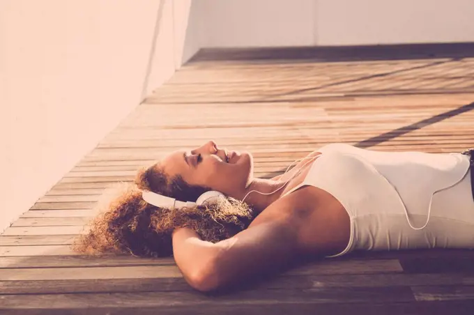 Young woman with eyes closed lying on wooden floor listening music with headphones