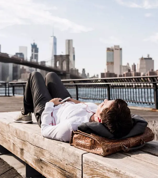 USA, Brooklyn, businesswoman lying on bench listening music with earphones