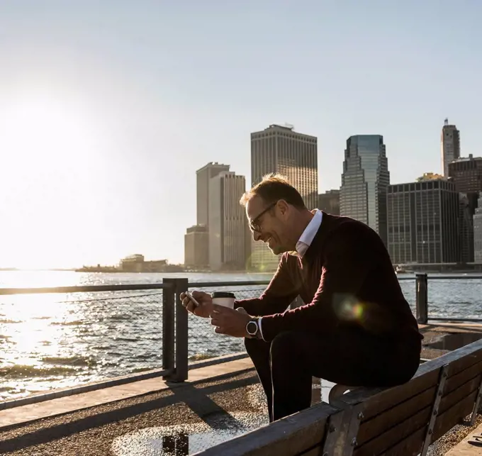 USA, Brooklyn, smiling man with coffee to go sitting on bench looking at smartphone